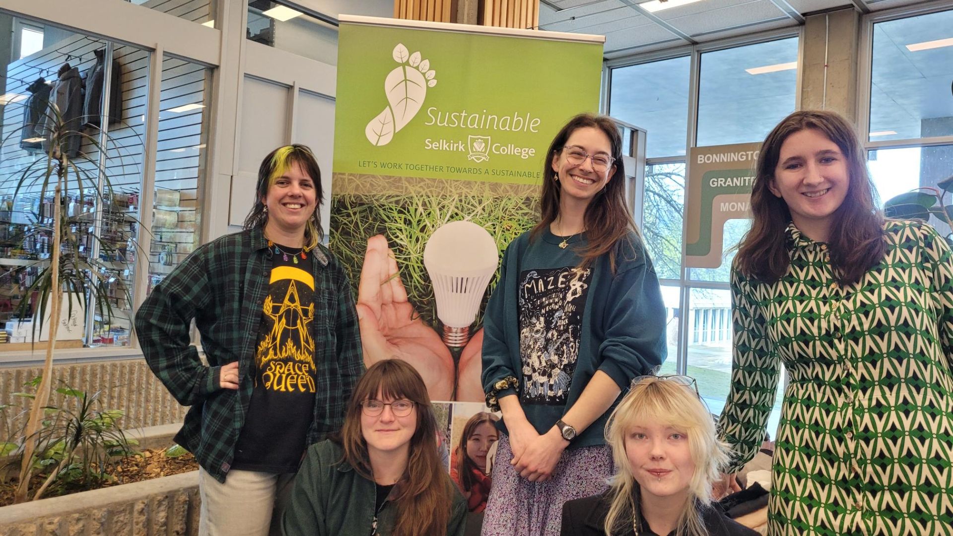 Four female Selkirk College students stand smiling before a Sustainable Selkirk banner