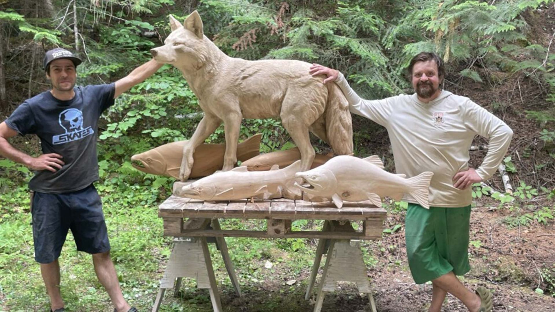 Carvers stand beside a wooden carving of a coyote and salmon