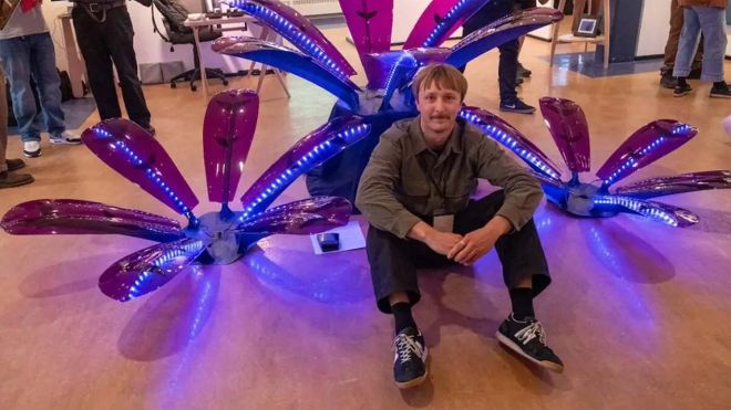 A student sits in front of a flower sculpture made of steel, purple acrylic and polycarbonate