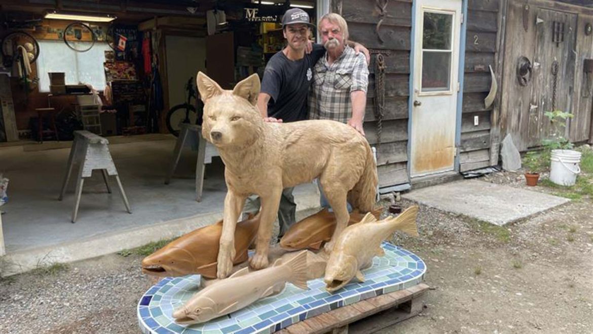 Two artists stand with a carving of a coyote