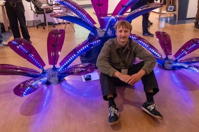 A student sits in front of a flower sculpture made of steel, purple acrylic and polycarbonate