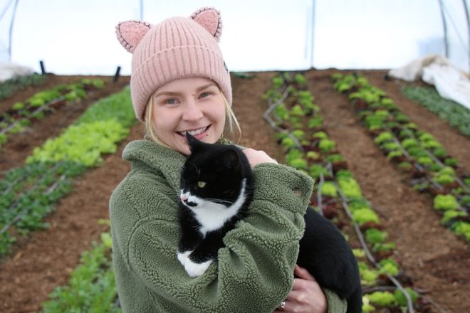 Sustainability Coordinator Kayla Tillapaugh stands in a greenhouse holding a cat and smiling